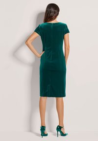 Eine Frau trägt ein knielanges, kurzärmeliges, smaragdgrünes Samtkleid mit einem Reißverschluss im Rücken, kombiniert mit passenden High Heels, und steht mit dem Gesicht zu einer Wand.