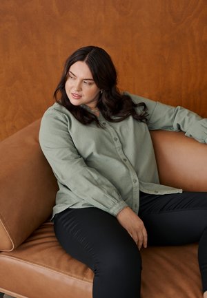 Femme aux cheveux ondulés foncés allongée sur un canapé en cuir marron, portant une chemise verte clair boutonnée et un pantalon noir, regardant vers sa gauche.