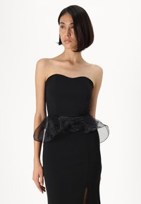 Vestido preto sem alças com corpete justo, babados transparentes esvoaçantes na cintura e uma fenda alta de um lado. Textura de tecido suave.