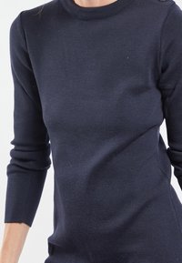 Femme portant un pull ajusté bleu marine foncé à manches longues avec un col rond et un détail de boutons à l'épaule, montré du cou aux hanches.