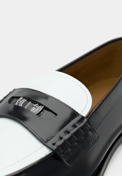 Μαύρο γυαλιστερό δερμάτινο μοκασίνι με λευκή δερμάτινη λεπτομέρεια, διακοσμημένη με κομμένο σχέδιο και ασημένια μεταλλικά στοιχεία. Το εσωτερικό έχει καφέ ταμπά φινίρισμα.