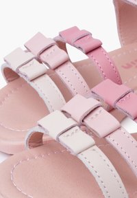 Sandalias planas de color rosa claro con múltiples tiras; material de cuero suave con hebillas cuadradas y un sutil detalle de costura en la suela.