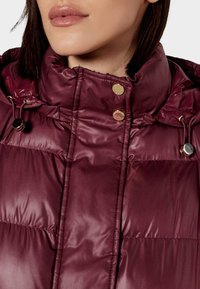 Giacca imbottita color bordeaux con colletto alto, caratterizzata da dettagli in metallo dorato, coulisse e texture trapuntata. Vista ravvicinata della scollatura.