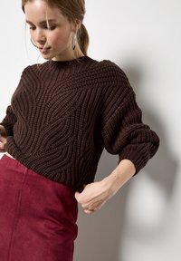 Femme ajustant un pull en tricot texturé marron foncé, portant de grandes boucles d'oreilles en créole et une jupe oversized rouge profond haute à la taille, le tout sur fond blanc.