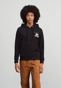 Fekete pamut kapucnis pulóver elülső zsebbel, húzózsinóros kapucnival és hímzett grafikai részlettel, amely egy karaktert ábrázol. A modellen barna nadrág van.