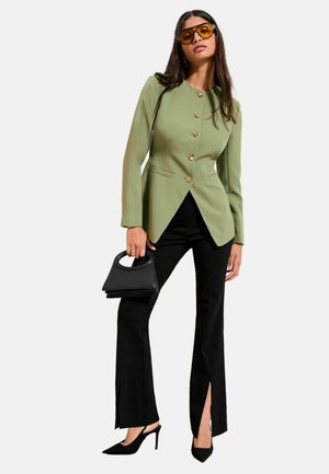 Green tailored blazer with gold buttons, paired with black flared trousers featuring side slits. Model holds small black handbag, wearing oversized sunglasses.