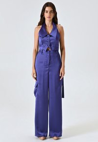 Completo in satin viola composto da due pezzi con top corto senza maniche, colletto, chiusura a bottoni e pantaloni a gamba larga con tasche.