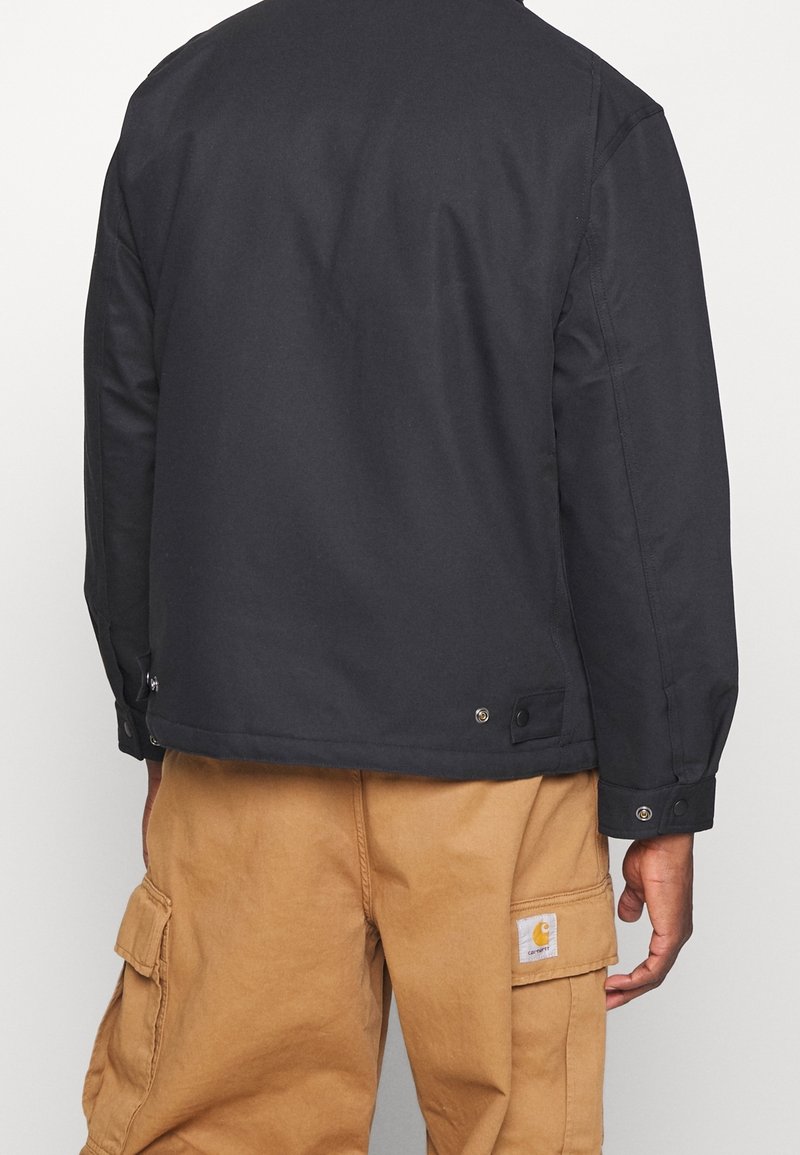 Černá bunda s přímým střihem, knoflíky na lemu a hladkou strukturou, doplněná khaki cargo kalhotami s kapsami.