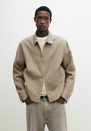 Giovane uomo in piedi contro uno sfondo neutro, indossa una giacca beige con cerniera, una camicia chiara sotto e pantaloni grigio chiaro con le mani in tasca.