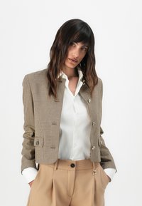 Barna kockás mintájú blézer rövid szabással, ezüst gombokkal és testhezálló fazonnal, fehér ingre húzva, bézs nadrággal párosítva.