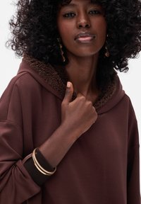Bruine capuchontrui met een pluche, donkerbruine fleece kraag. Heeft een oversized ontwerp en opgerolde mouwen. Draag het met gouden armbanden.