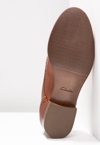 Semelle de chaussure en cuir marron avec un fond texturé en caoutchouc et des coutures détaillées le long des bords. Présente le logo "Clarks" en relief.