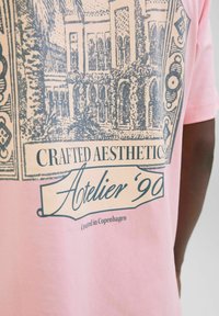 Lyserød bomulds t-shirt med et detaljeret blåt grafisk print af en bygning og blade, med teksten "Crafted Aesthetics" og "Atelier '90".