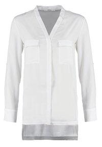 Vit långärmad blus med V-hals, två bröstfickor och ett genomskinligt bottenlager. Tillverkad av lätt, slätt tyg.