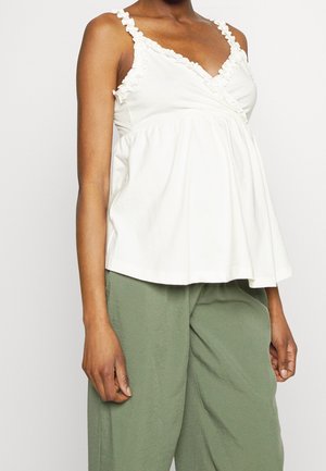 Biały, bezrękawnik z falbankowymi ramiączkami, owijanym fasonem i rozszerzanym dołem. Połączony z luźnymi, jasnozielonymi spodniami dla swobodnego wyglądu.