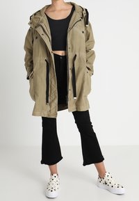 Khaki nylon parka with a hood, two front pockets, black cord accents, paired with cropped black top, black flared jeans, and white polka dot sneakers.