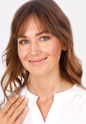Gold necklace with a delicate chain and small oval pendant, worn with a white blouse. Model has long brown hair and a neutral makeup look.