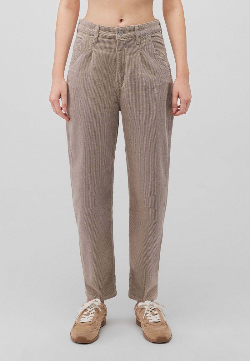 Personne portant un pantalon en velours côtelé beige taille haute avec poches avant et bouton, associé à des baskets marron clair avec lacets blancs, fond uni.