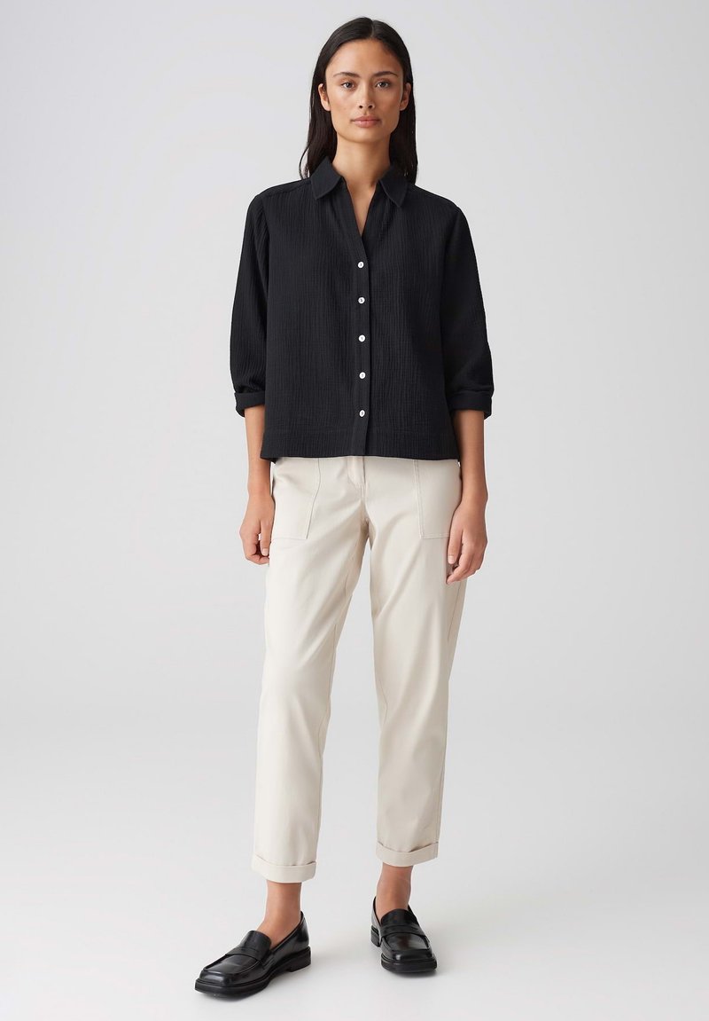 Zwarte button-up shirt met een gestructureerde stof, opgerolde mouwen en witte knopen, gecombineerd met lichtbeige broek en zwarte loafers.