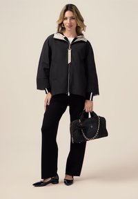 Veste noire à capuche avec zip, pantalon noir et ballerines noires brillantes. Tenant un sac noir avec une chaîne en accent. Textures lisses dans l'ensemble.