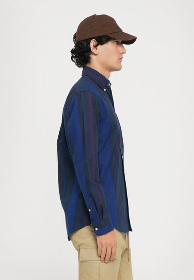 Camisa de botones a rayas azules con acentos verdes y rojos, combinada con pantalones cargo beige, llevada con una gorra de béisbol marrón.