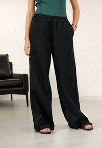 Sorte, løstsiddende bukser med elastisk taljebånd og sidelommer. Letvægtsstof med en glat tekstur. Båret med flade, tanfarvede sandaler.