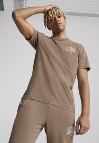 Brun bomulds T-shirt med korte ærmer og PUMA-logo på brystet, parret med matchende brune joggers med lignende branding.