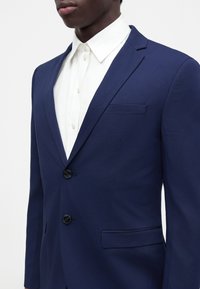 Uomo che indossa un blazer blu navy aderente con due bottoni neri sopra una camicia bianca abbottonata, ritagliato sotto il naso.