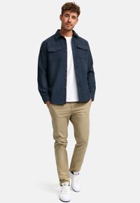 Camisa de botones azul marino con dos bolsillos en el pecho, forro interior gris, combinada con pantalones beige y zapatillas blancas. Textura de tela suave.