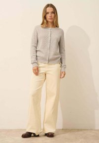 Cardigan en maille gris clair avec un motif texturé, présentant une fermeture à boutons et des bords festonnés, associé à un pantalon en velours côtelé crème à jambes larges.