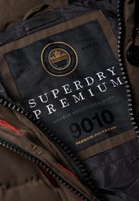 Brun og sort jakkeindvendig etiket med "Superdry Premium"-logo, tekst med hvide og gyldne accenter samt angivelse af premium andedun.