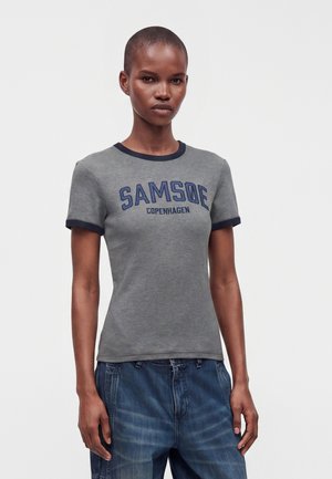 Femme portant un t-shirt gris ajusté avec bordure bleu marine et texte "Samsøe Copenhagen", associé à un jean bleu ample sur un fond uni.