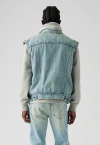 Šviesiai mėlyna džinsinė liemenė dėvima ant pilkos gaubtuotos megztinės. Turi padraskytas kraštines, spaudėmis užsegamus sagčius ir šiek tiek trumpintą kirpimą. Derinama su išblukusiais mėlynais džinsais.