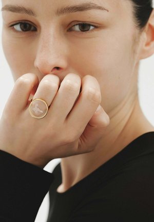 Primer plano de una persona con expresión neutral que lleva un anillo de oro con un pequeño diseño de mariposa en la mano levantada.