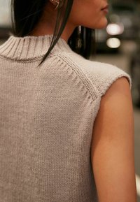 Szary sweter bez rękawów wykonany na drutach z prążkowanym kołnierzem, o gładkiej teksturze i czystych liniach. Widoczne detale szwów na ramionach.