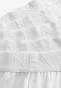 Biały bawełniany pas pojedyńczo z wytłoczonym logo "NEXT", charakteryzujący się gładką fakturą i miękkim, elastycznym fasonem, ułożony w stos.