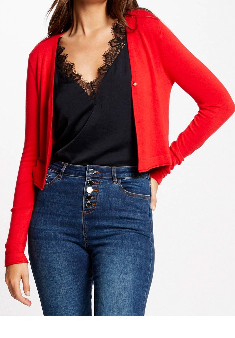 Femme portant un cardigan court rouge sur un haut noir avec bordure en dentelle, associé à un jean bleu taille haute avec plusieurs boutons.