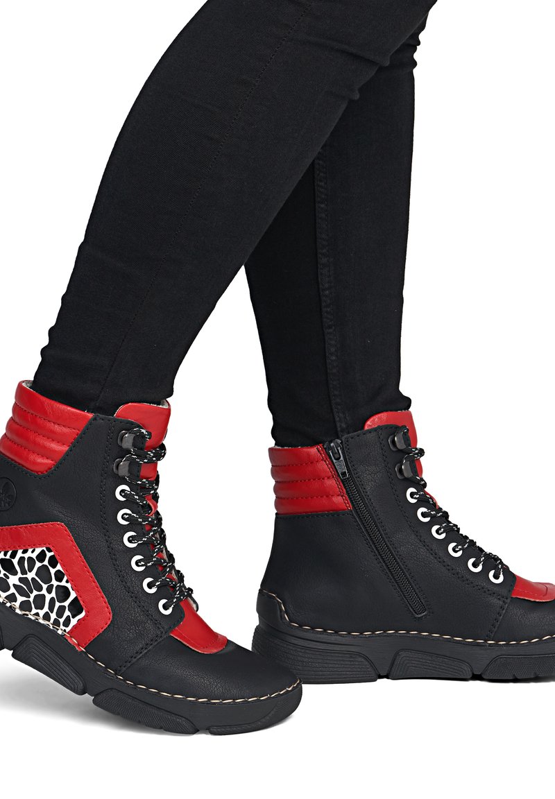 Bottes hautes noires et rouges avec lacets, fermeture éclair latérale et détail imprimé animal, portées avec un pantalon noir ajusté.