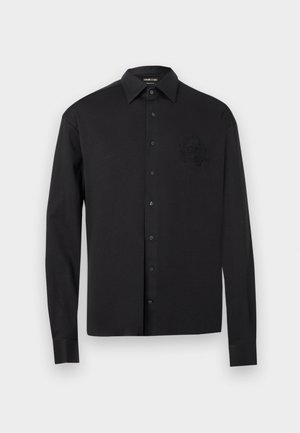 Chemise noire à manches longues avec boutons, col pointu et blason brodé subtil sur le côté gauche de la poitrine, présentée sur un fond blanc.