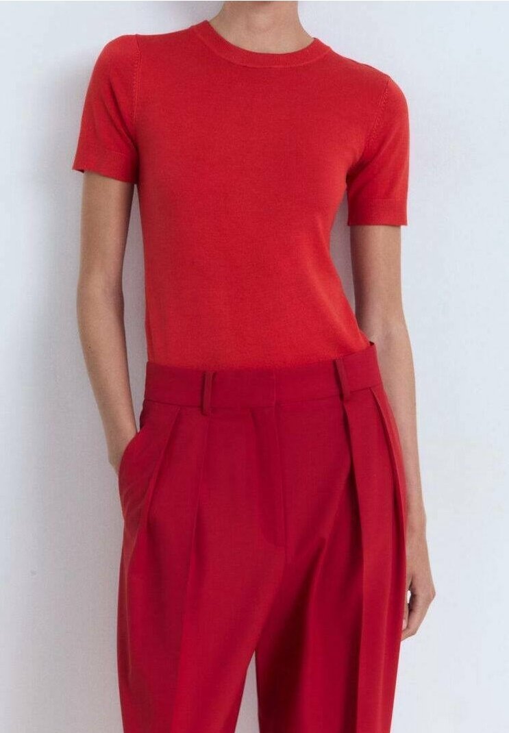 Femme portant un haut rouge ajusté à manches courtes rentré dans un pantalon rouge plissé taille haute, mains dans les poches, sur fond blanc uni.