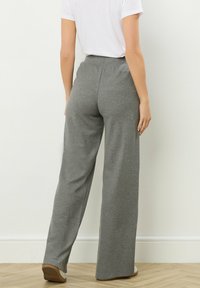 Persoon in hoge taille, wijde grijze textuurbroek en een witte kortlopende shirt, staand tegen een witte muur.