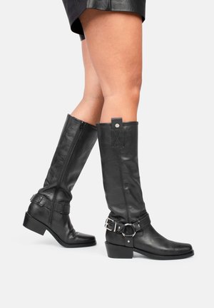 Botas de cuero negras hasta la rodilla con cierre de cremallera, que presentan una correa ancha y hebilla a lo largo del tobillo. Diseño de puntera cuadrada y tacón block.