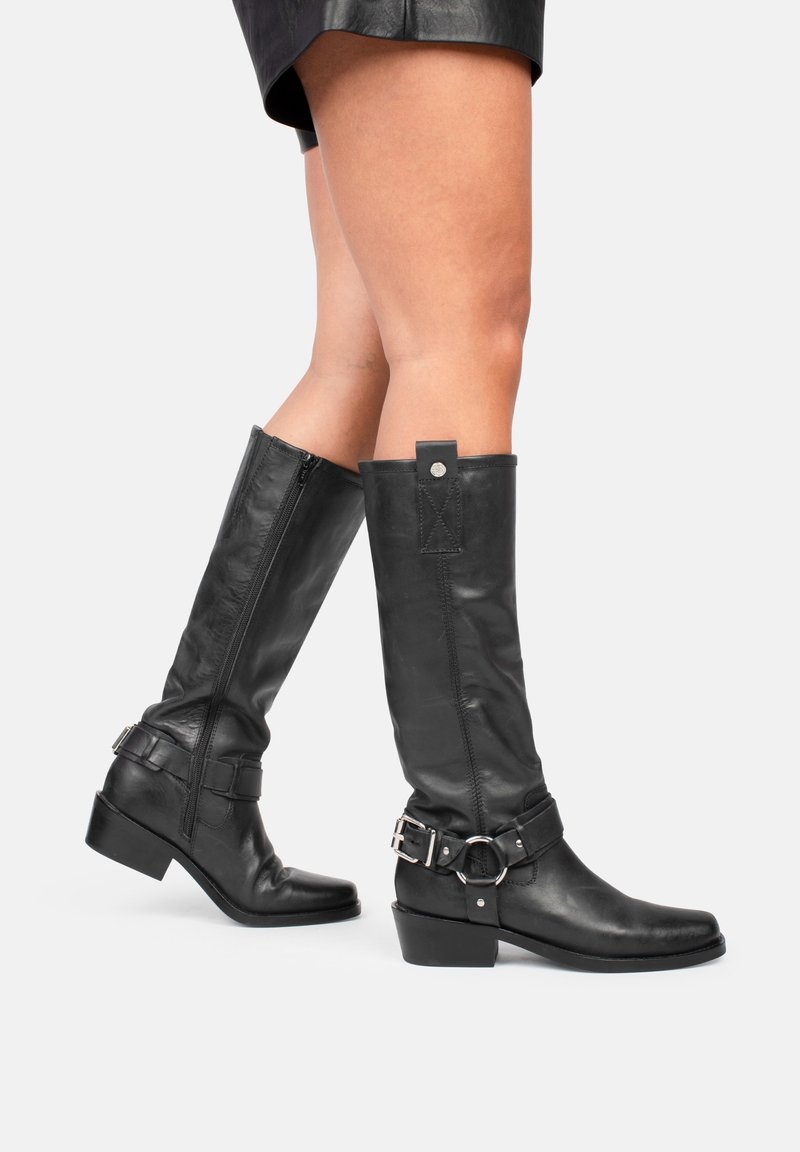 Botas de cuero negras hasta la rodilla con cierre de cremallera, que presentan una correa ancha y hebilla a lo largo del tobillo. Diseño de puntera cuadrada y tacón block.
