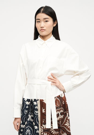 Γυναίκα με μακριά μαύρα μαλλιά, που φορά λευκό πουκάμισο με ζώνη και κουμπιά, καθώς και φούστα με σχέδιο σε ναυτικό μπλε, μπεζ και καφέ αποχρώσεις με λουλουδένιο μοτίβο.