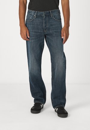 Blauwe spijkerbroek met een ontspannen pasvorm, voorzien van vijf zakken, contrasterende stiksels en een knoopsluiting. Draagt het met zwarte sneakers.