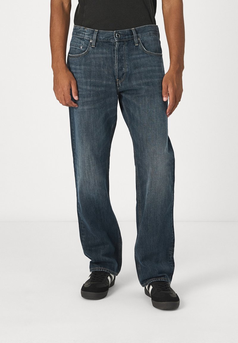 Jeans de mezclilla azul con un corte relajado, que cuentan con cinco bolsillos, costuras en contraste y un cierre de botón. Combinados con zapatillas negras.