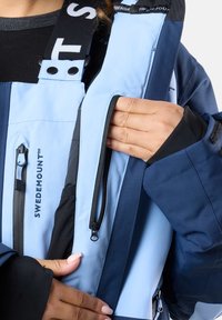 Niebieska i granatowa wodoodporna kurtka z zamkami błyskawicznymi oraz regulowanymi paskami. Posiada kieszeń na rękę i elastyczne mankiety dla lepszego dopasowania. Widoczne logo SWEDEMOUNT.