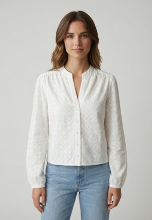 Mujer con cabello castaño ondulado, vestida con una blusa blanca de manga larga con botones y patrón floral texturizado, y jeans azul claro, posando frente a un fondo gris.