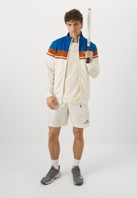 Chaqueta blanca con cremallera y rayas azules y marrones, camiseta beige, pantalones cortos blancos y zapatillas grises. El individuo sostiene un bate.