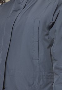 Námořnická modrá voděodolná bunda s jemnou strukturou, vybavená zipem a boční kapsou na levé straně, navržená pro funkčnost.